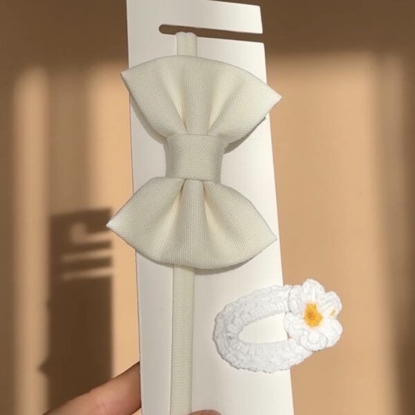 Set de lux pentru bebeluși conținând o bentiță albă cu fundiță și o agrafă croșetată cu floare, prezentate pe un suport alb.