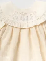 Detaliu guler rochiță fetițe ivoriu cu broderie florală delicată în nuanțe pastelate și volănaș fin pe fundal alb neutru.