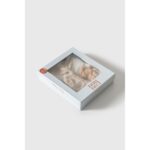 Set cadou premium pentru bebeluși în cutie albă cu fereastră, conținând hăinuțe din in bej și accesorii asortate, prezentat pe fundal gri deschis.