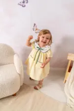 Fetiță zâmbitoare purtând o rochiță galben pal cu guler brodat cu lămâi și fundiță verde, pozând într-un decor luminos cu fluturi și fotoliu pufos.