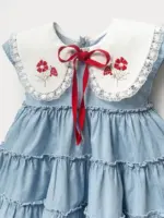 Rochiță fetițe din denim bleu cu guler alb supradimensionat, broderie cu flori roșii și căpșuni, finisată cu dantelă și fundiță roșie contrastantă.
