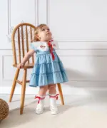Fetiță mică purtând o rochiță din denim bleu cu guler alb brodat și fundă roșie, stând lângă un scaun de lemn, pe un fundal clasic alb.