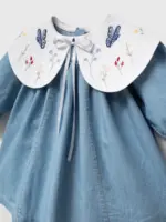 Detaliu pe gulerul alb brodat cu fluturi albaștri și flori multicolore al unei rochițe pentru fetițe din denim moale.